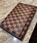 A rectangular wooden cutting board with a checkered pattern of light and dark brown wood squares, shown resting on a speckled granite countertop.