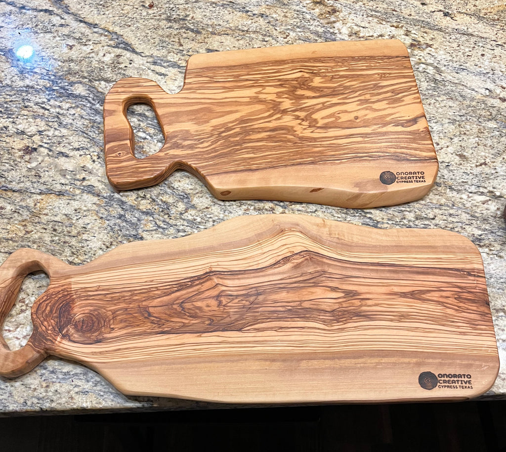 Two handcrafted olive wood cutting boards with loop handles resting on a granite kitchen countertop, with a person's shoes and legs visible at the bottom edge.