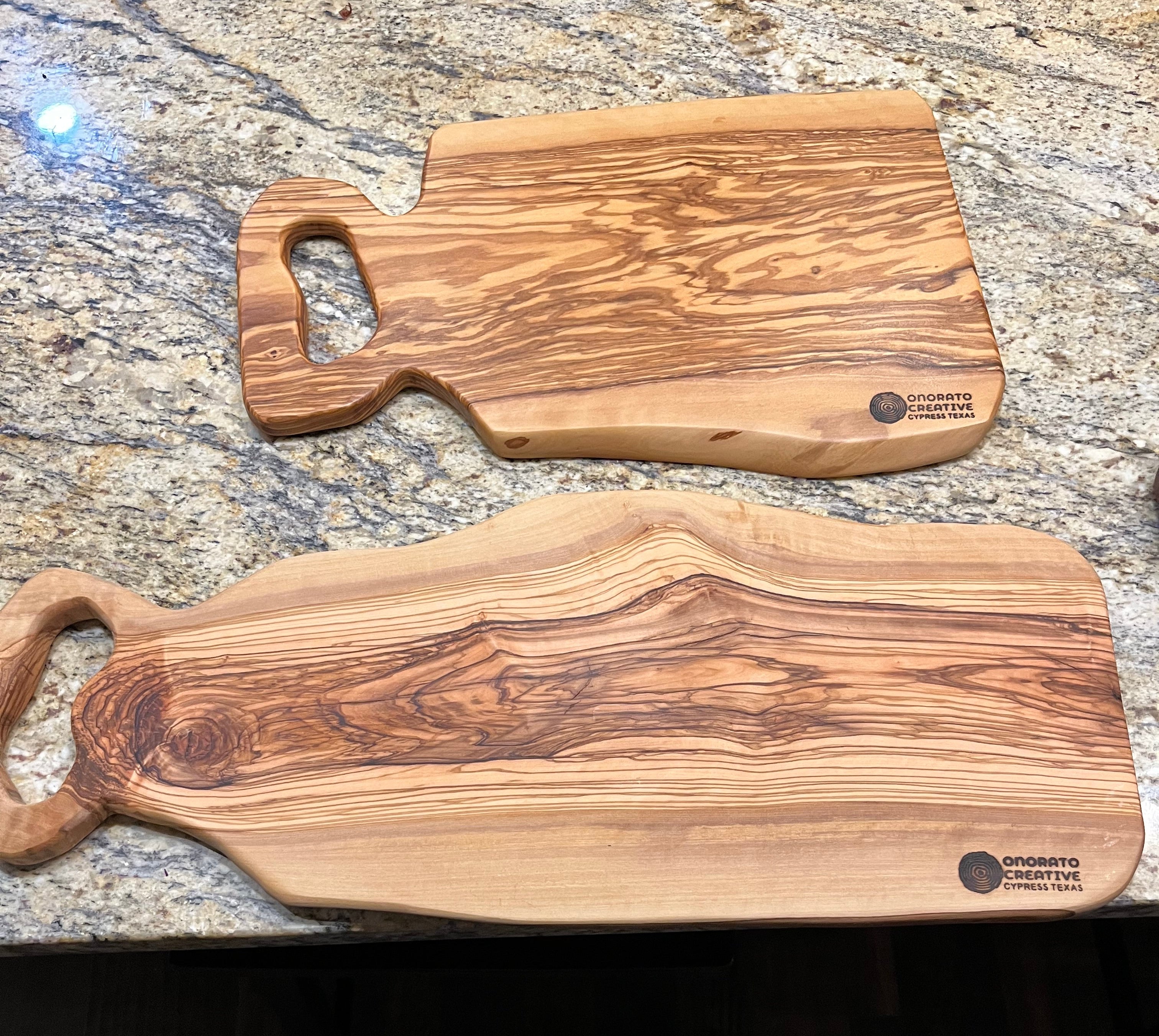 Two handcrafted olive wood cutting boards with loop handles resting on a granite kitchen countertop, with a person's shoes and legs visible at the bottom edge.