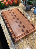 A rectangular wooden cutting board with a geometric inlay pattern, resting on a granite kitchen countertop.